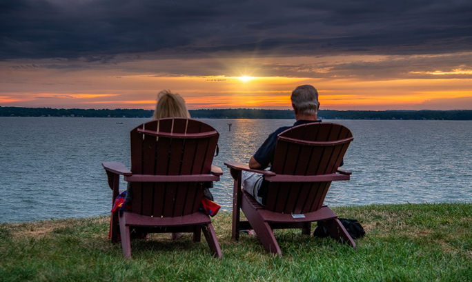 A couple sitting in Adirondack chairs watch a beautiful sunset over the lake.